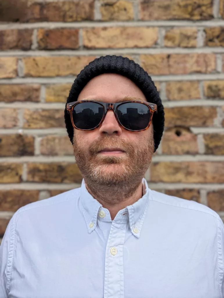 Jay Springett wearing a black beanie, tortoiseshell sunglasses, and a white button-down shirt against a brick wall.