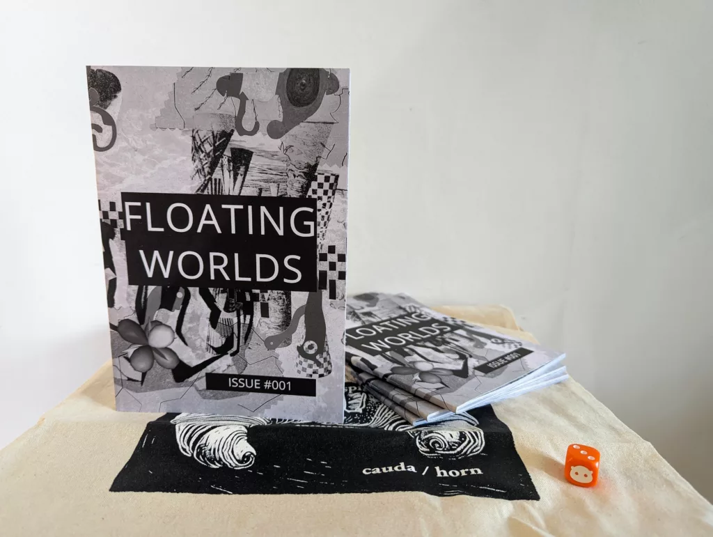 Monochrome zine Floating Worlds Issue #001 with an abstract collage cover, standing on a tote bag beside a stack of copies and a small orange die.
